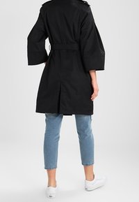 Černý trenčkot s širokými rukávy, zúženým pasem a rozparkem na zadní straně. Vyznačuje se hladkým povrchem a minimem kovových detailů.