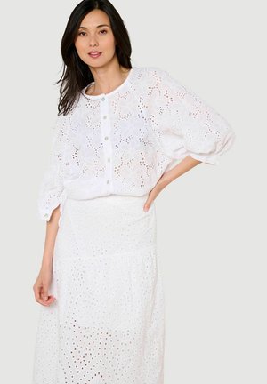 Mujer que lleva una blusa blanca de encaje ojete con botones y una falda blanca de encaje ojete a juego, de pie con una mano en la cadera.