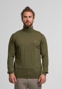 Olivgrüner Rollkragenpullover aus Strickstoff, mit langen Ärmeln und gerippten Bündchen, sowie einem kleinen Logo auf der Brust.