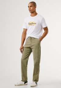 Homem vestindo uma t-shirt branca da Pepe Jeans London, calças verde-oliva e ténis brancos com detalhes verdes, de pé com uma mão no bolso.