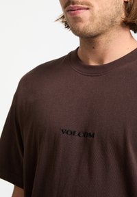 T-shirt en coton marron avec des manches courtes, un col rond et un logo « VOLCOM » brodé en noir centré sur la poitrine. Texture lisse.