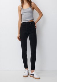 Jeans skinny negros de cintura alta con una textura suave, que cuentan con cinco bolsillos y un cierre de botón, combinados con una camiseta sin mangas gris y zapatillas blancas.