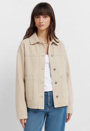 Kobieta ubrana w beżową, zapinaną na guziki kurtkę z dużymi przednimi kieszeniami, na białej bluzce i niebieskich dżinsach, stojąca na jasnym, jednolitym tle.