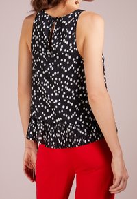 Top senza maniche nero con pois bianchi, orlo plissettato e dettaglio a chiave sulla schiena. Abbinato a pantaloni rossi. Tessuto dalla superficie liscia.