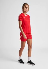 Tenue athlétique rouge comprenant une chemise à manches courtes et un short assorti, fabriquée en matériau léger, avec des accents blancs en forme de chevron sur le short.