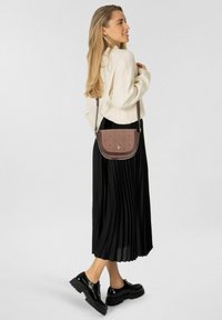 Braune Leder-Umhängetasche mit strukturiertem Muster, abgerundetem Überschlag und verstellbarem Riemen. Getragen zu einem cremefarbenen Pullover und einem schwarzen plissierten Rock.