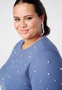 Blauwe trui van zacht materiaal, versierd met verspreide parelaccenten. Ronde halslijn en aansluitende mouwen. Oorbellen met hangende parels.