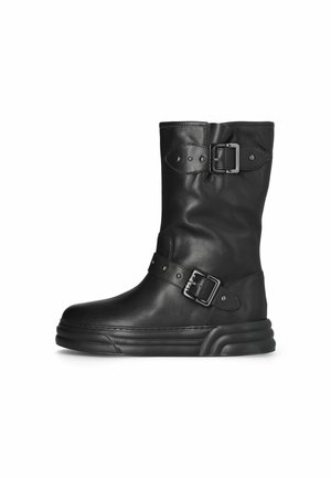 Botte en cuir noire mi-mollet avec semelle épaisse, deux sangles ajustables à boucle et bout arrondi orienté vers la gauche sur fond blanc.