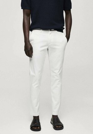 Tummaihoinen henkilö, joka pukeutuneena valkoisiin kapeisiin housuihin, mustiin sandaaleihin ja lyhythihaiseen mustaan teksturoituun toppiin, yksiväristä taustaa vasten.