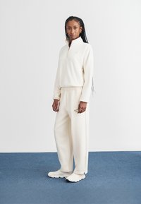 Cream fleece pullover with half-zip and logo; matching cream wide-leg sweatpants with an elastic waist; paired with light-colored sneakers.