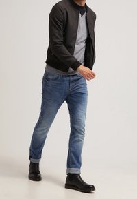 Veste bomber noire, pull gris, jean bleu et bottines noires. Le jean est légèrement délavé avec des revers, la veste a une texture lisse.