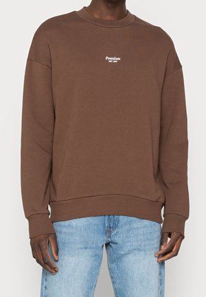 Person trägt einen braunen Pullover mit "Premium EST. 1990" Text vorne, kombiniert mit hellblauen Jeans, stehend vor einem einfarbigen Hintergrund.
