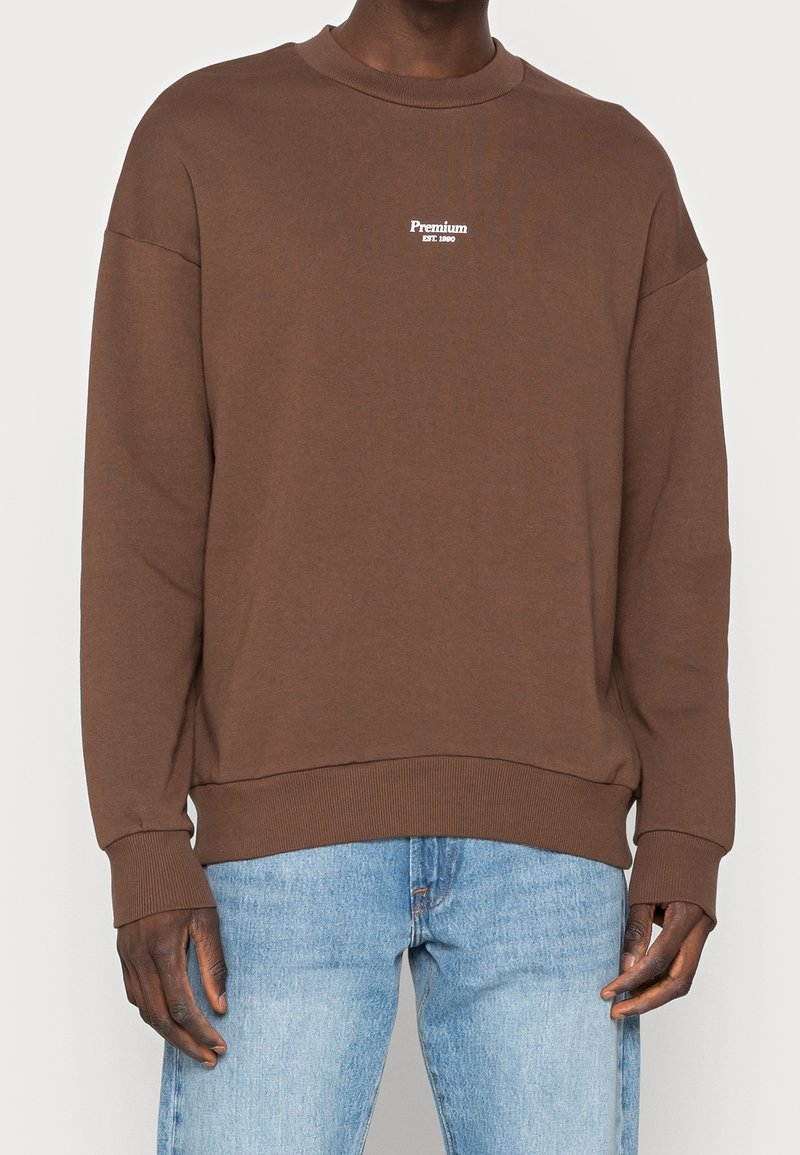 Personne portant un sweat marron avec le texte "Premium EST. 1990" sur le devant, associé à un jean bleu clair, debout sur un fond uni.