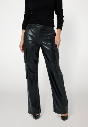 Person trägt schwarze Cargo-Hose aus Kunstleder mit Seitentaschen, schwarzes Langarmoberteil und schwarze spitz zulaufende Schuhe vor einem einfarbigen Hintergrund.