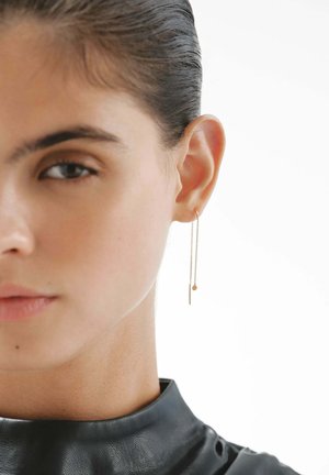 Pendientes de cadena dorada que presentan una barra delgada y una pequeña gota circular, colocados elegantemente en la oreja del modelo contra un cuello negro.