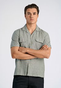 Camisa de lino de manga corta, de color verde claro, con dos bolsillos en el pecho y detalles de botones, que lleva puesto un hombre con los brazos cruzados sobre un fondo sencillo.
