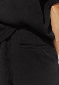 Gros plan d'une personne portant un pantalon noir texturé avec une poche passepoilée arrière et un haut noir assorti, montrant une partie de son bras.