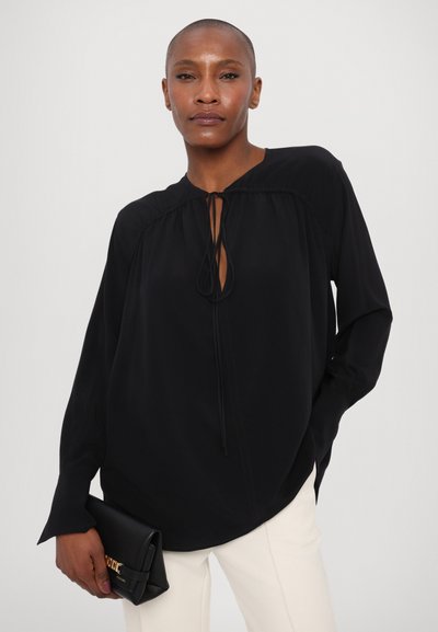 Blusa nera a maniche lunghe con scollatura rotonda, dettaglio con fiocco sul davanti e leggere arricciature sulle spalle. Abbinata a una pochette nera.