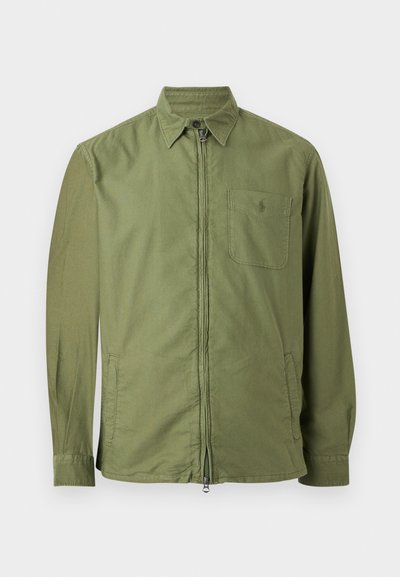 Olíva zöld hosszú ujjú dzseki elülső cipzárral, gombos gallérral, mellkasi zsebbel és két oldalzsebbel, fehér sík háttéren bemutatva.