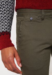 Olivgrüne Hose mit einer Seitentasche, Knopfverschluss mit einem Herzdetail und einer glatten, strukturierten Oberfläche. Der Pullover hat geometrische Muster.