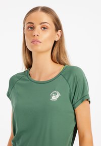 Camisa verde de manga corta con cuello redondo y un estampado de logo en el pecho, hecha de tejido suave y transpirable con una textura lisa.
