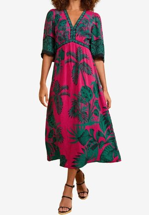 Femme portant une robe midi rose avec des motifs de feuilles tropicales vertes et des sandales à bride de cheville noires, se tenant devant un fond uni.