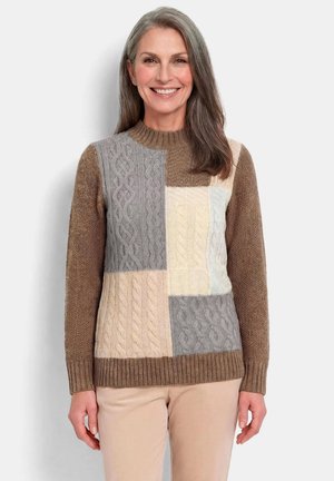 Frau mit langen grauen Haaren lächelt, trägt einen Patchwork-Zopfmuster-Pullover in Braun-, Grau-, Creme- und Beigetönen sowie beige Hosen.