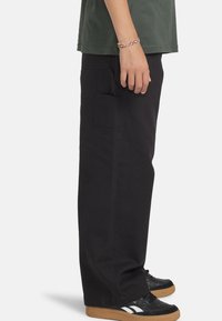 Zwarte wijde pantalon gemaakt van duurzaam materiaal. Bevat een zijzak en is gecombineerd met zwarte sneakers en een groene blouse.