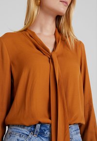 Rustrød bluse med knyttet hals, blød tekstur og løs pasform. Sammen med lyseblå denim, der fremhæver blusens fluiditet.