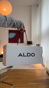 Biała torba zakupowa ALDO z czarnym logo, trzymana w dłoni. Torba ma solidną konstrukcję i czarne papierowe uchwyty. Tło zawiera źródła światła.