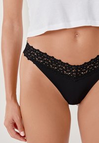 Black lace-trimmed thong featuring a floral pattern at the waistband, made from a smooth fabric with a sleek design.