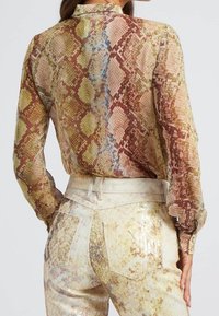 Blouse transparente en imprimé python avec manches longues, aux couleurs marron, vert et bleu. Associée à un pantalon à taille haute texturé et orné de sequins.