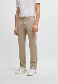 Beige chino nadrág, slim fit szabással, gombos záródással, elülső zsebekkel rendelkezik, és sima pamut keverékből készült. Fehér sportcipővel párosítva.