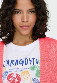 Coral knit cardigan over a white t-shirt featuring colorful seafood graphics and text. Straight hair, neutral expression.