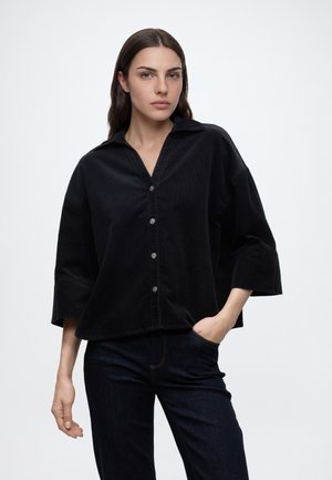 Femme aux longs cheveux foncés portant une chemise noire ample boutonnée et un pantalon foncé, debout avec une main dans la poche sur un fond uni.