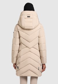 Beige puffer jacket with a chevron pattern, padded design, and a large hood. Features include black side accents and a tag near the hem.