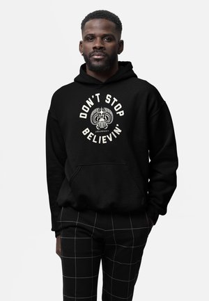 Muž s vousy, oblečený v černé mikině s nápisem "Don't Stop Believin' Journey" a designem skaraba, doplněný černobílými kostkovanými kalhotami.
