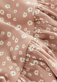 Tissu rose clair avec une texture côtelée, orné de fleurs blanches à centres marron dispersées. Le motif est doux et fluide avec des plis.