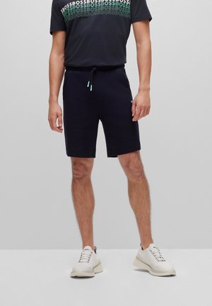 Mann trägt schwarze Shorts, schwarzes T-Shirt mit wiederholtem "BOSS"-Text und weiße Sneaker, steht vor einem schlichten grauen Hintergrund.