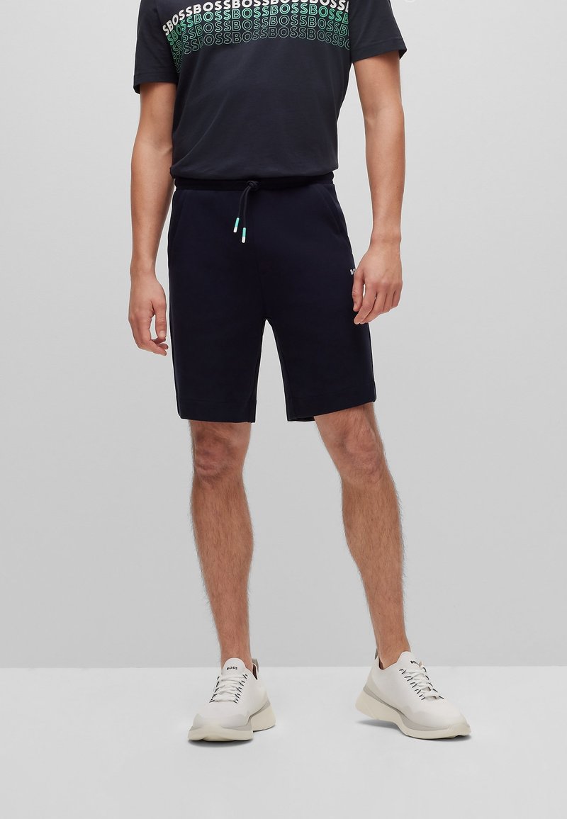 Homme portant un short noir, un T-shirt noir avec le texte répétitif "BOSS" et des baskets blanches, debout sur un fond gris uni.