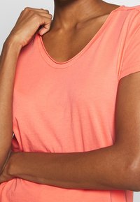 Korálové tričko s krátkým rukávem vyrobené z měkké tkaniny, které má kulatý výstřih a volný střih s hladkým povrchem a čistými švy.