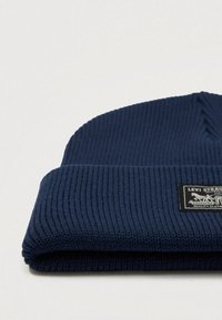 Navy blue ribbed knit beanie with a folded cuff and a prominent black label featuring design details and text.