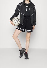 Chaqueta deportiva negra con acentos blancos, camiseta negra, pantalones cortos negros con rayas blancas, zapatos grises y negros, sosteniendo un balón de baloncesto.