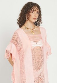 Mujer con el pelo rizado lleva una prenda rosa claro con flecos sobre un top a juego desgarrado y un collar de oro con dijes.