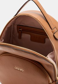 Zaino in pelle marrone di forma rotonda, con zip dorata, tasca interna e etichetta con logo. La texture è granulosa, con fodera liscia.