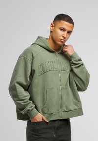 Maslinasto zelena zip-up dukserica s utisnutim logom "KARL KANI", oversized kroj, veliki prednji džepovi, rebrasti završeci rukava i donjeg dela, materijal od pamučne mešavine.