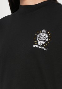 T-shirt nera con colletto alto, caratterizzata da un elaborato design ricamato a forma di cuore con un serpente, un occhio e il testo "JUST CAVALLI" in bianco.