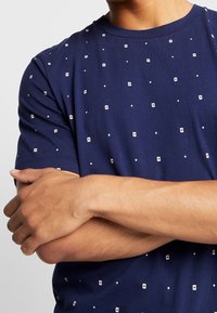 Man crossing arms wearing a navy blue T-shirt with a white dotted square pattern on a plain background.