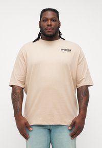 Beżowy t-shirt z krótkim rękawem o luźnym kroju. Posiada małe czarne logo "Tropica" na lewej piersi. Miękki, gładki materiał bawełniany.