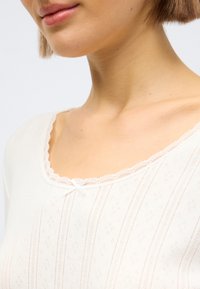 Λευκή μπλούζα με κυματιστό λαιμόκοψη, διακοσμημένη με δαντέλα και ένα μικρό φιόγκο. Το ύφασμα έχει υφή με κάθετες ρίγες και μικρά διαμάντια.